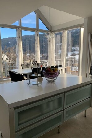 A modern kitchen with a large island, a dining area with a table and chairs, and a view of a mountainous landscape through large windows. - Haus Kreischberg by Interhome (Sankt Georgen am Kreischberg)