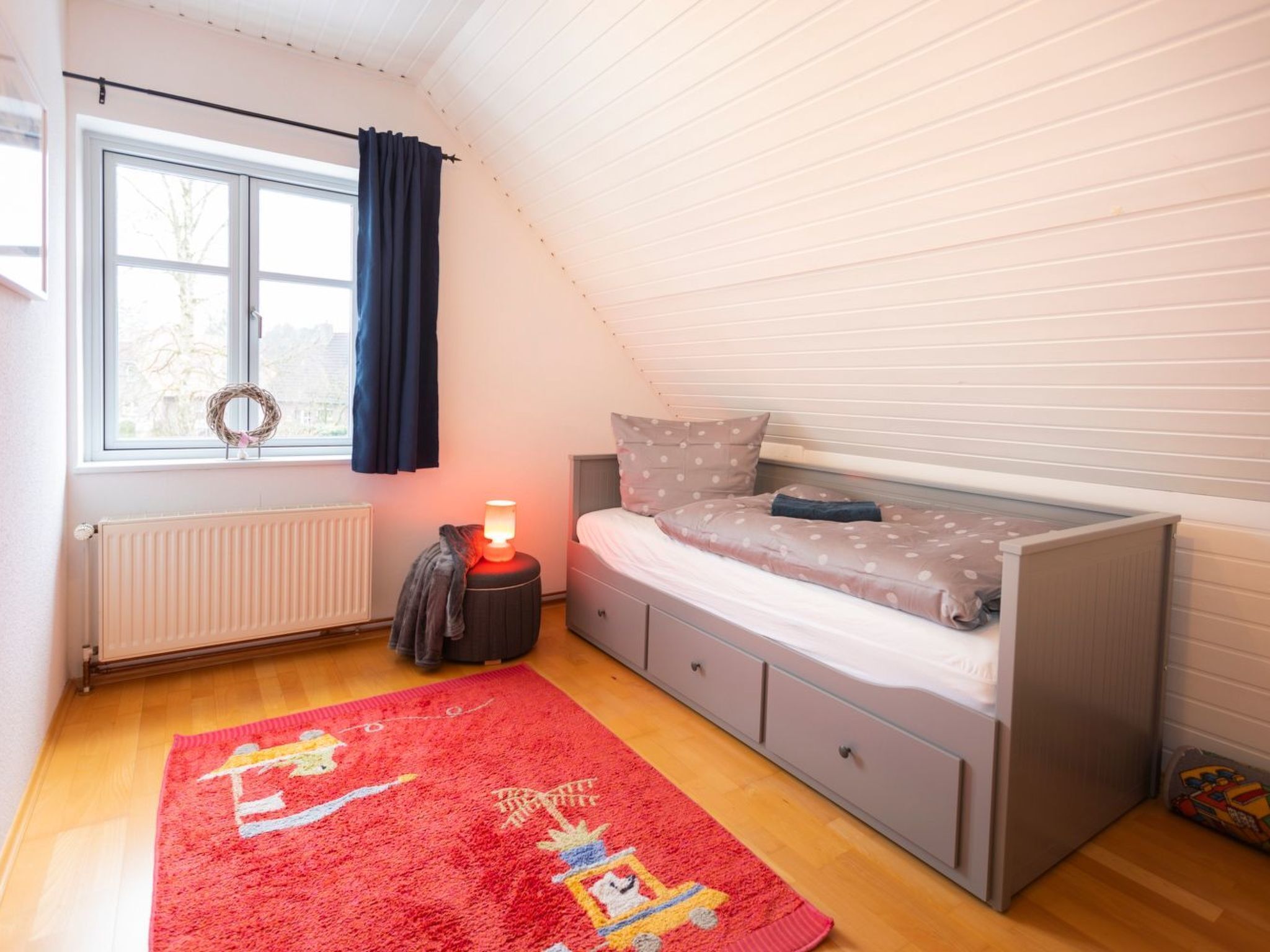 A bedroom with a bed, a window with curtains, a radiator, a lamp, and a red rug.