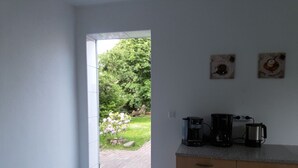 Uma cozinha com cafeteira, chaleira e bancada. Há uma porta que dá para um jardim com gramado e árvore.