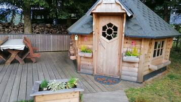 Una casita de juegos de madera con un ventanal hexagonal, una puerta y un pequeño porche con plantas.