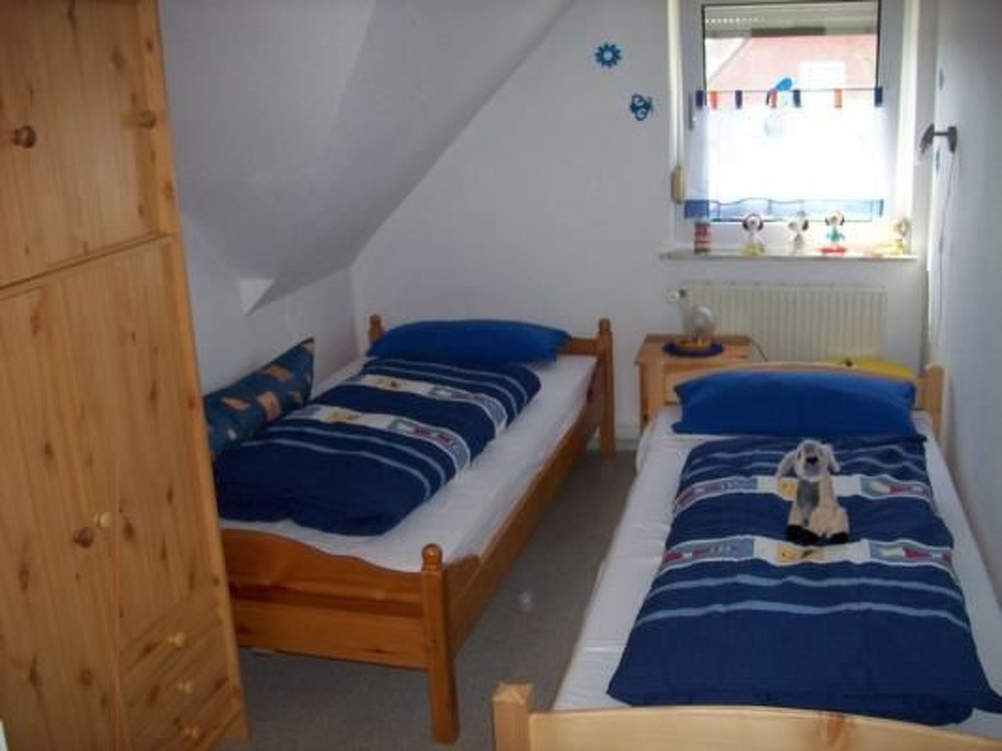Ein kleines, gemütliches Schlafzimmer mit zwei Holzbetten, jeweils mit blauer Bettwäsche und Kissen. Links befindet sich ein Holzschrank und rechts ein kleiner Tisch mit einer Lampe.