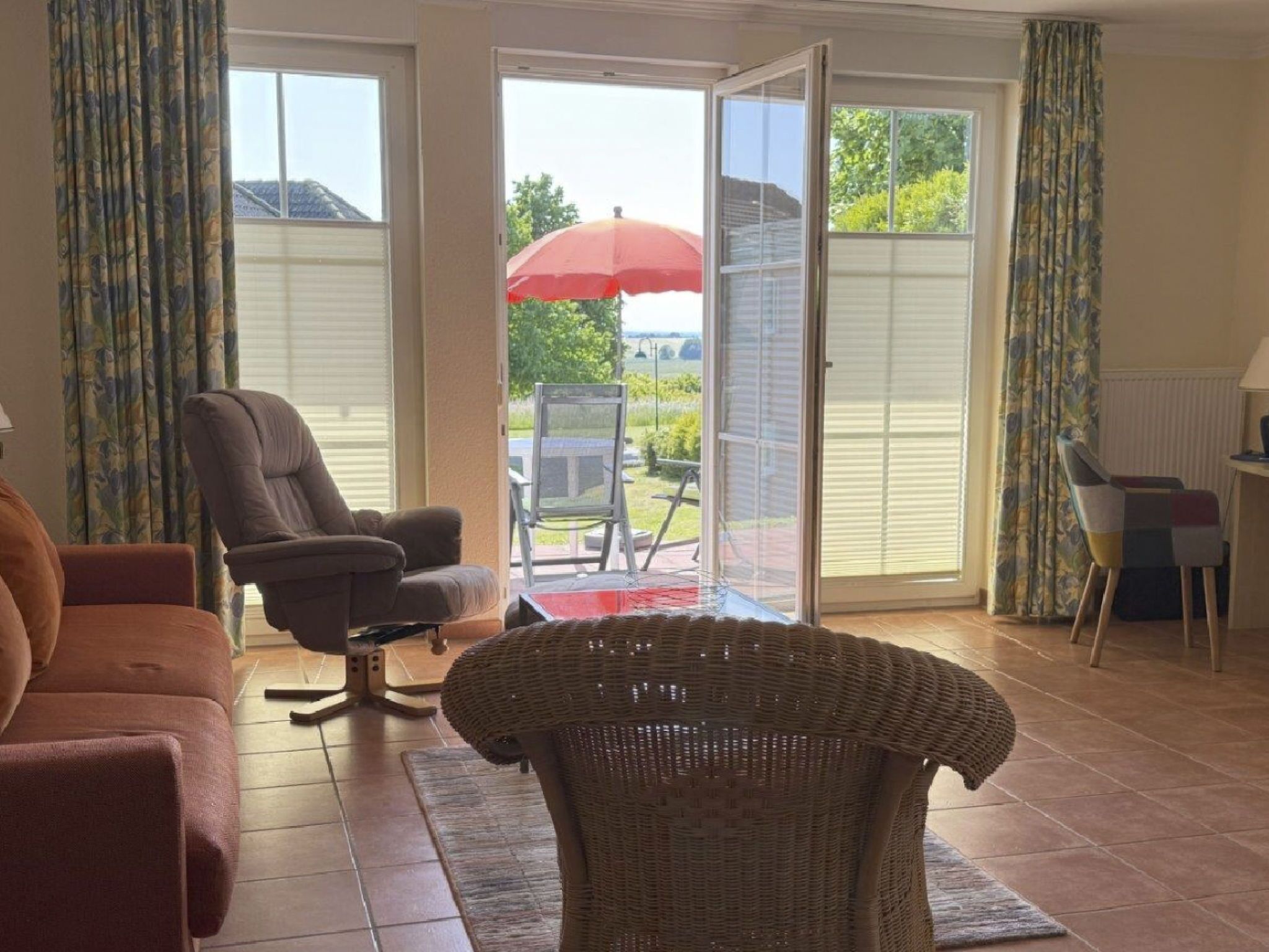 Een woonkamer met een bank, fauteuil en rieten stoel. Er is een schuifdeur naar een buitenruimte met een tafel en stoelen.