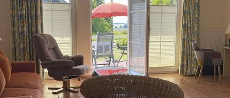 Een woonkamer met een bank, fauteuil en rieten stoel. Er is een schuifdeur naar een buitenruimte met een tafel en stoelen.