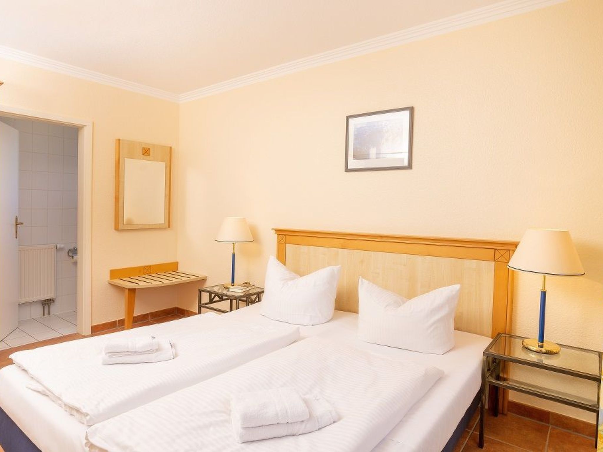 Habitación de hotel con dos camas, cabecero de madera, una mesita de noche con lámpara, un cuadro enmarcado en la pared y un baño visible a través de una puerta abierta.
