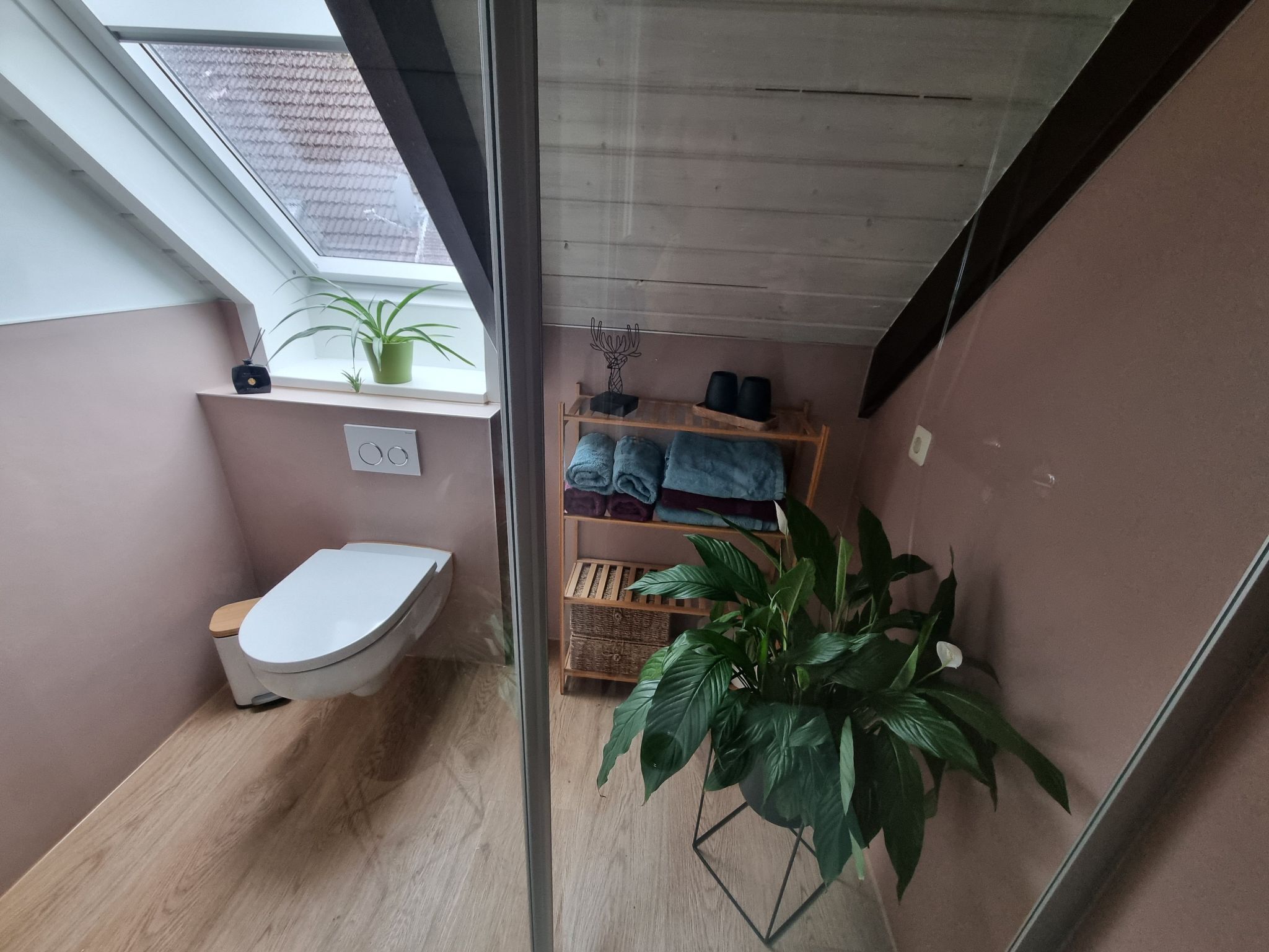 Un baño pequeño con inodoro, estante con toallas, planta en maceta y ventana.
