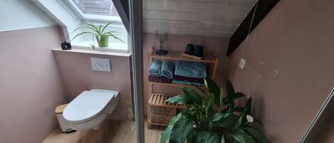 Un baño pequeño con inodoro, estante con toallas, planta en maceta y ventana.