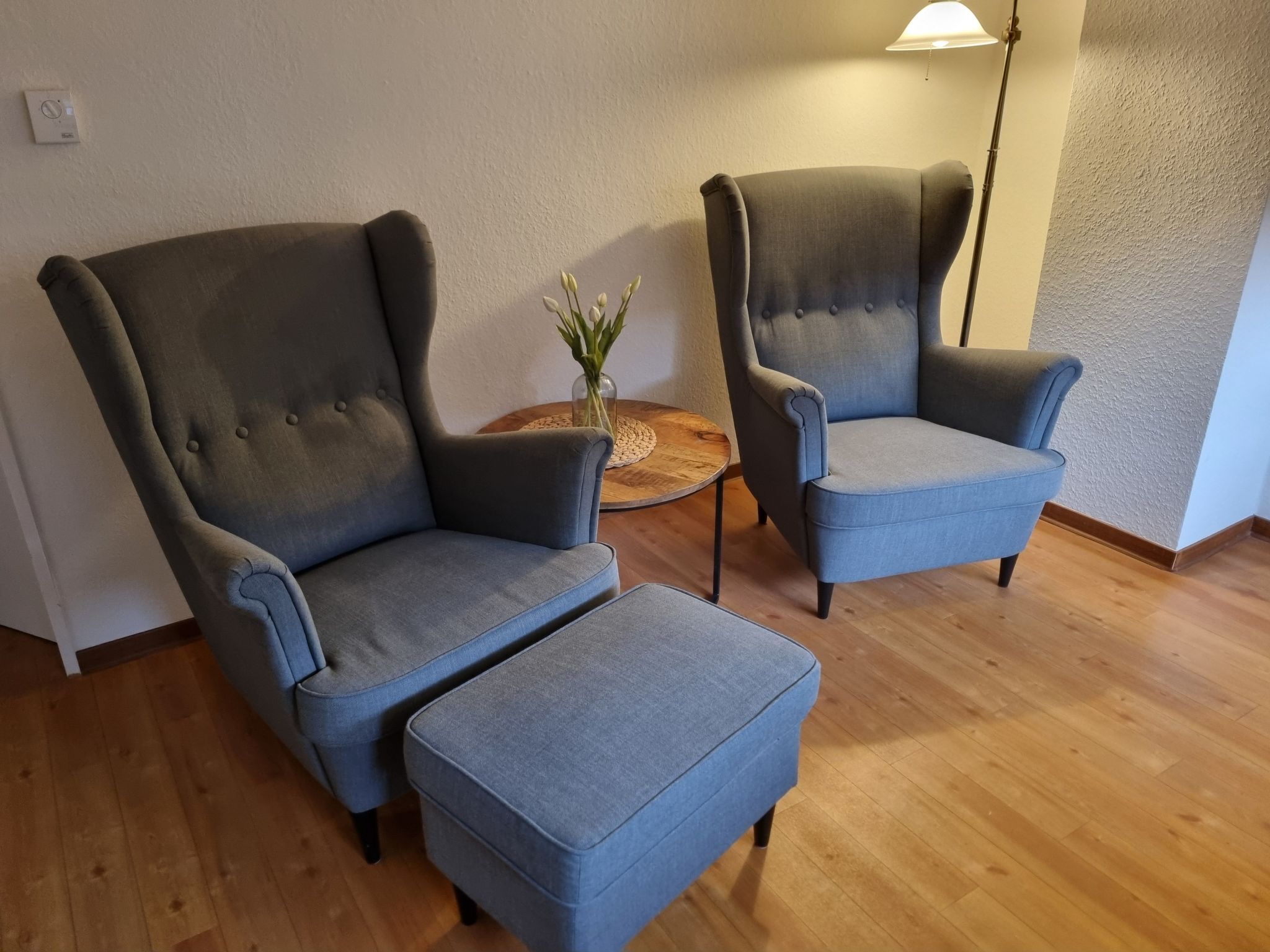 Deux fauteuils gris assortis d’un repose-pieds, une petite table ronde en bois surmontée d’un vase de fleurs et une lampe sur pied dans une pièce au plancher en bois et aux murs texturés.