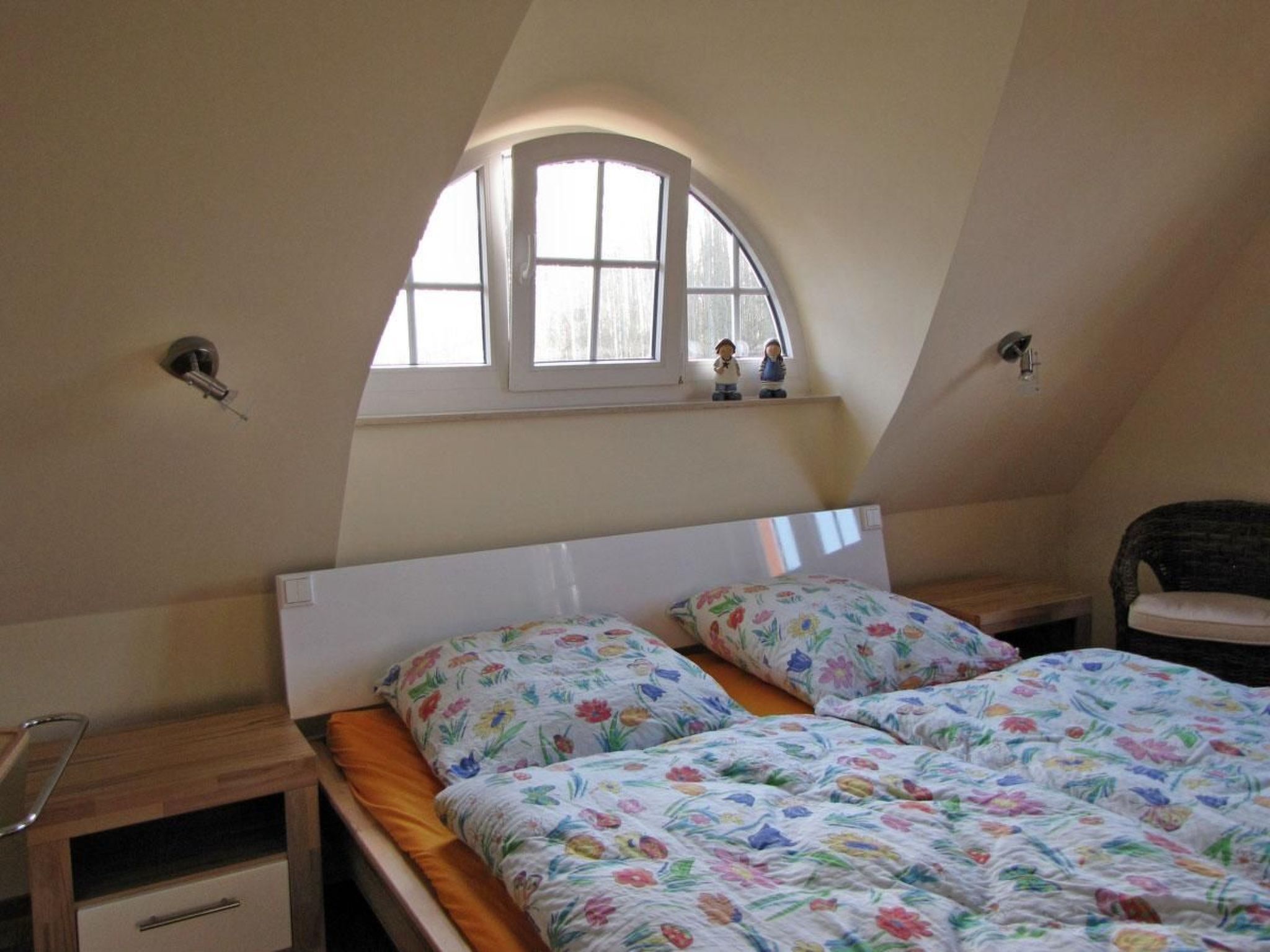 A bedroom with a sloped ceiling, a window with two figurines, a bed with floral bedding, and a nightstand with a lamp.