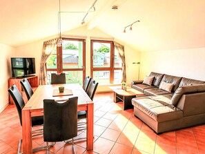 A dining area with a table and chairs, a sofa, a TV, and large windows with curtains. - Zeesenhaus 6 by Interhome (Ostseebad Sellin)