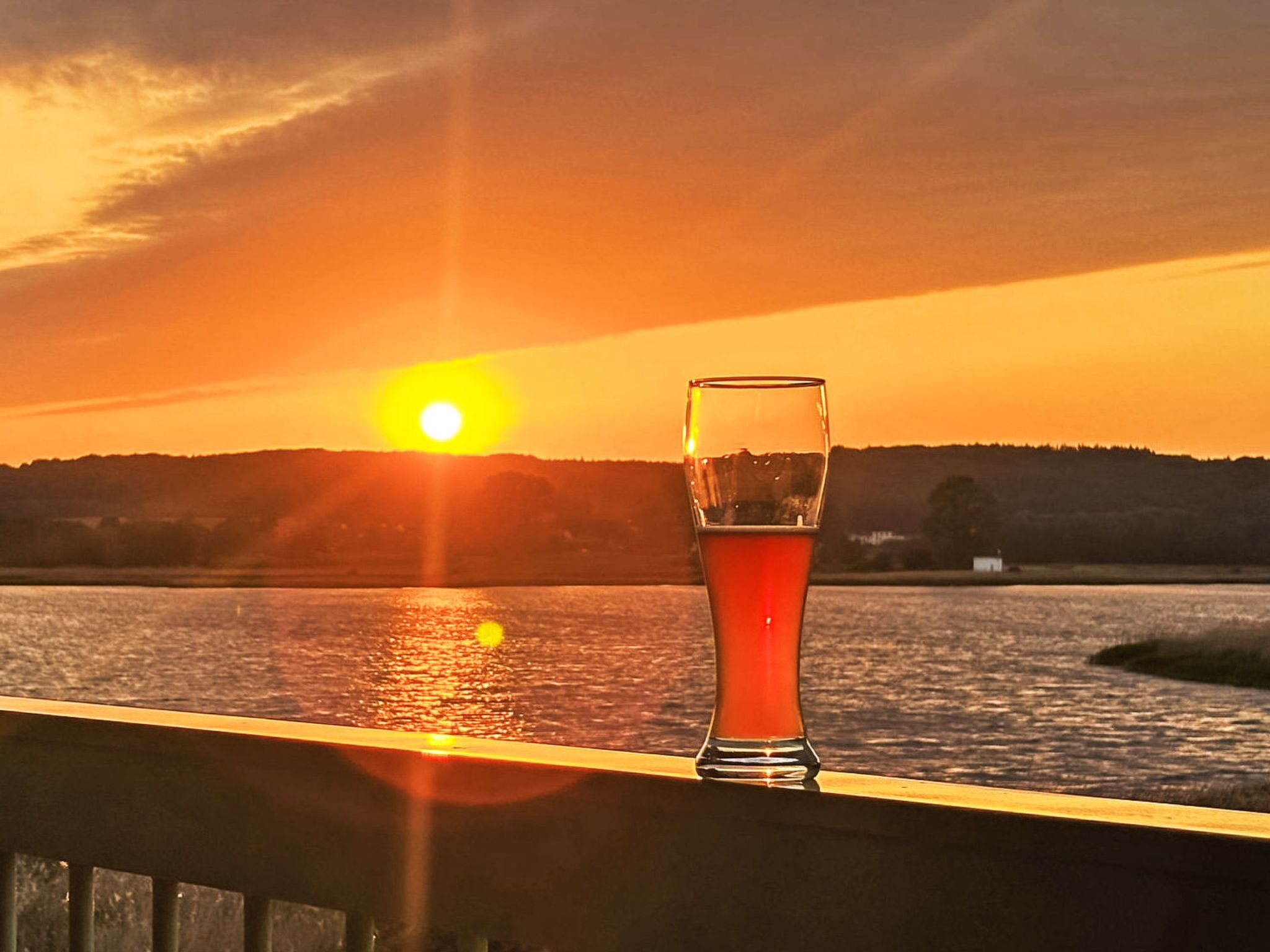 Tramonto su un corpo d'acqua con un bicchiere di liquido rosso su una ringhiera.