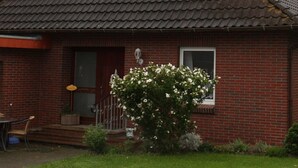 Une maison de plain-pied en briques, avec un toit en tuiles, une porte d’entrée et un petit porche agrémenté d’une chaise et d’un pot de fleurs.