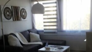 Un dormitorio moderno con cama, sofá, mesita de café y escritorio. Hay ventanas con persianas, un ventilador de pared y elementos decorativos en la pared.
