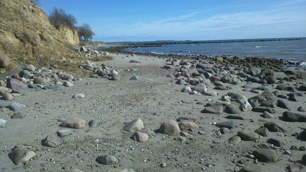 Ein felsiger Strand mit einer Klippe links und ruhigem Wasser im Hintergrund.