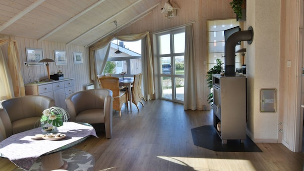 Ein großzügiger Wohnbereich mit Esstisch, Stühlen und einer Couch. Eine Küchenzeile mit Herd und Schränken ist ebenfalls vorhanden. Große Fenster sorgen für viel Tageslicht.