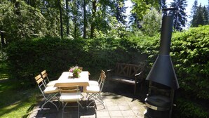 An outdoor patio with a table and chairs, surrounded by lush greenery and a chimney-style barbecue grill. - Gretelhaus by Interhome (Auerbach)