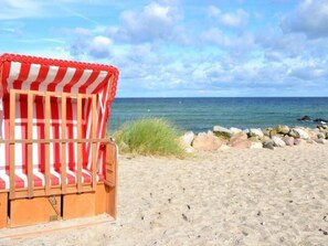 Une chaise de plage recouverte d’une toile rayée rouge et blanche, placée sur une plage de sable, avec une côte rocheuse et l’océan en arrière-plan.