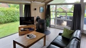 A modern living room with a wooden coffee table, a black leather sofa, a television, and a large window offering a view of the garden and outdoor furniture. - Suennhuus by Interhome (Brodersby)