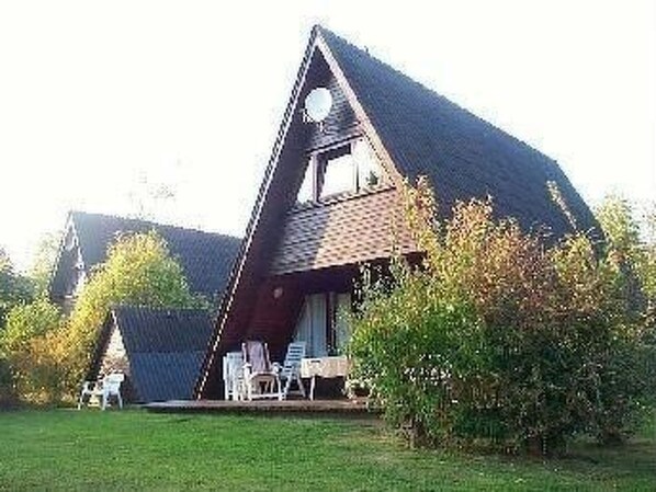 A triangular-shaped building with a sloped roof, a porch with chairs, and a grassy yard. - Spitzdachferienhaus mit Schwedenofen und Garten by Interhome (Damp)