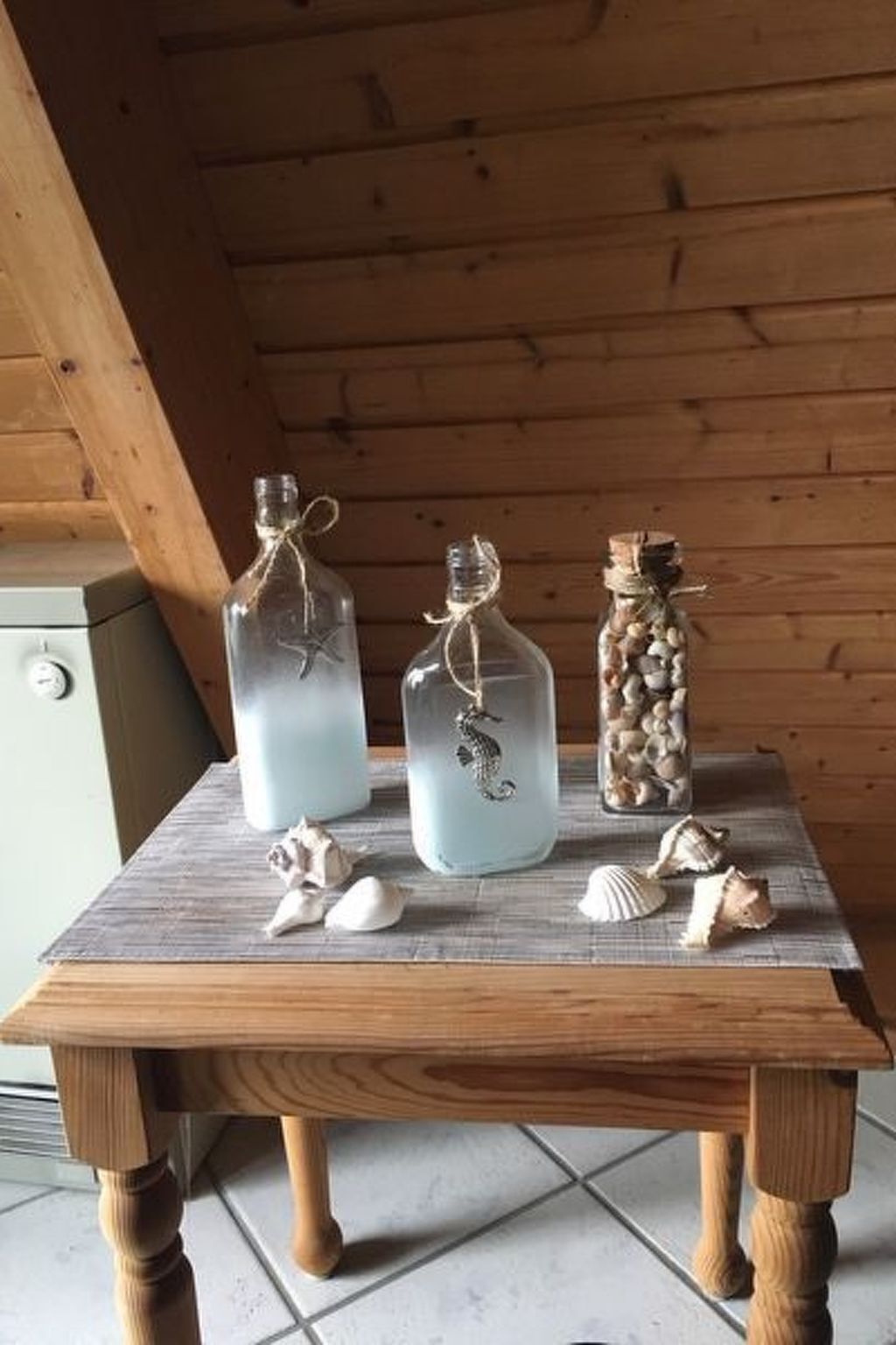 A wooden table with three glass bottles, one with a seahorse decoration, and a jar filled with seashells.