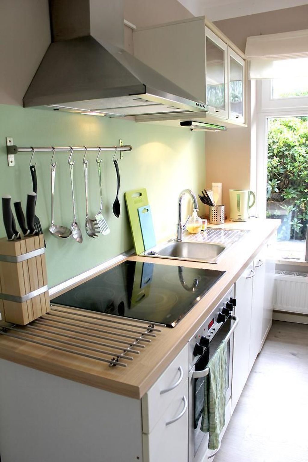 A modern kitchen with a built-in stove, a hanging utensil rack, a sink, and a window with a view of greenery.