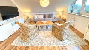 Un salon moderne doté d’un téléviseur à écran plat, d’un canapé, de deux fauteuils en osier, d’une table basse et d’une grande fenêtre offrant une vue dégagée.