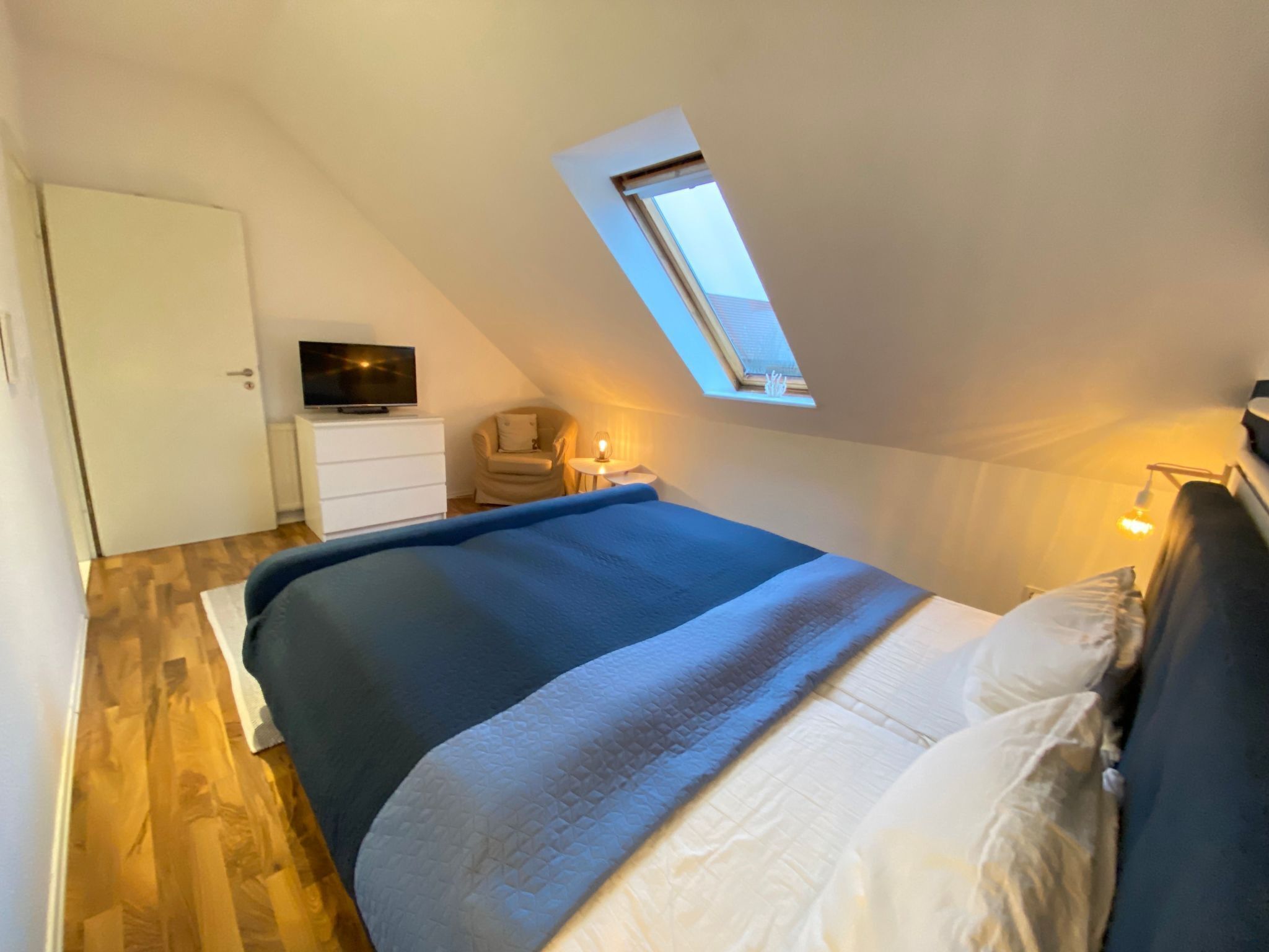 Une chambre à coucher avec un lit, un téléviseur posé sur une commode, une chaise, une petite table avec une lampe et un puits de lumière.