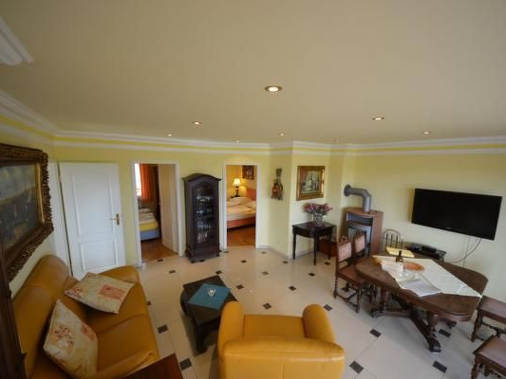 Quarto de hotel com sofá amarelo, uma mesa de jantar com cadeiras, televisão presa à parede e uma cama ao fundo.