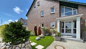 Ein zweistöckiges Backsteinhhaus mit Veranda, Kiesweg und einem kleinen Garten mit Steinen und Pflanzen.