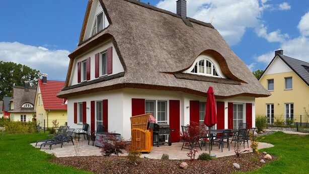 Ein traditionelles Haus mit einem Strohdach, roten Fensterläden und einer kleinen Außenbereich-Sitzgelegenheit mit Stühlen und einem Tisch.