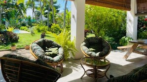 A room with two wicker chairs and a small round table, large windows with bamboo blinds, and a view of a lush garden with palm trees. - in Roches Noires mit Großem Garten by Interhome (Cap Malheureux)