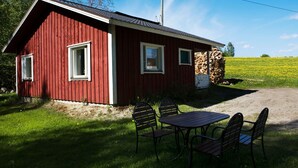 En röd trästuga med två fönster, ett bord och fyra stolar, placerad på en gräsyta.