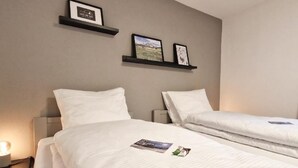 Hotellihuone, jossa on kaksi sänkyä, valkoiset vuodevaatteet ja yöpöytä, jossa on lamppu. Sänkyjen yläpuolella seinällä on kehystettyjä kuvia.