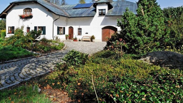 A white building with a sloped roof, a stone pathway, and a garden with various plants and trees.