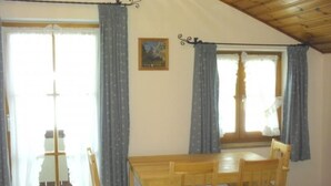 Een kamer met een houten tafel, vier stoelen, een raam met gordijnen en een klein schilderij aan de muur.