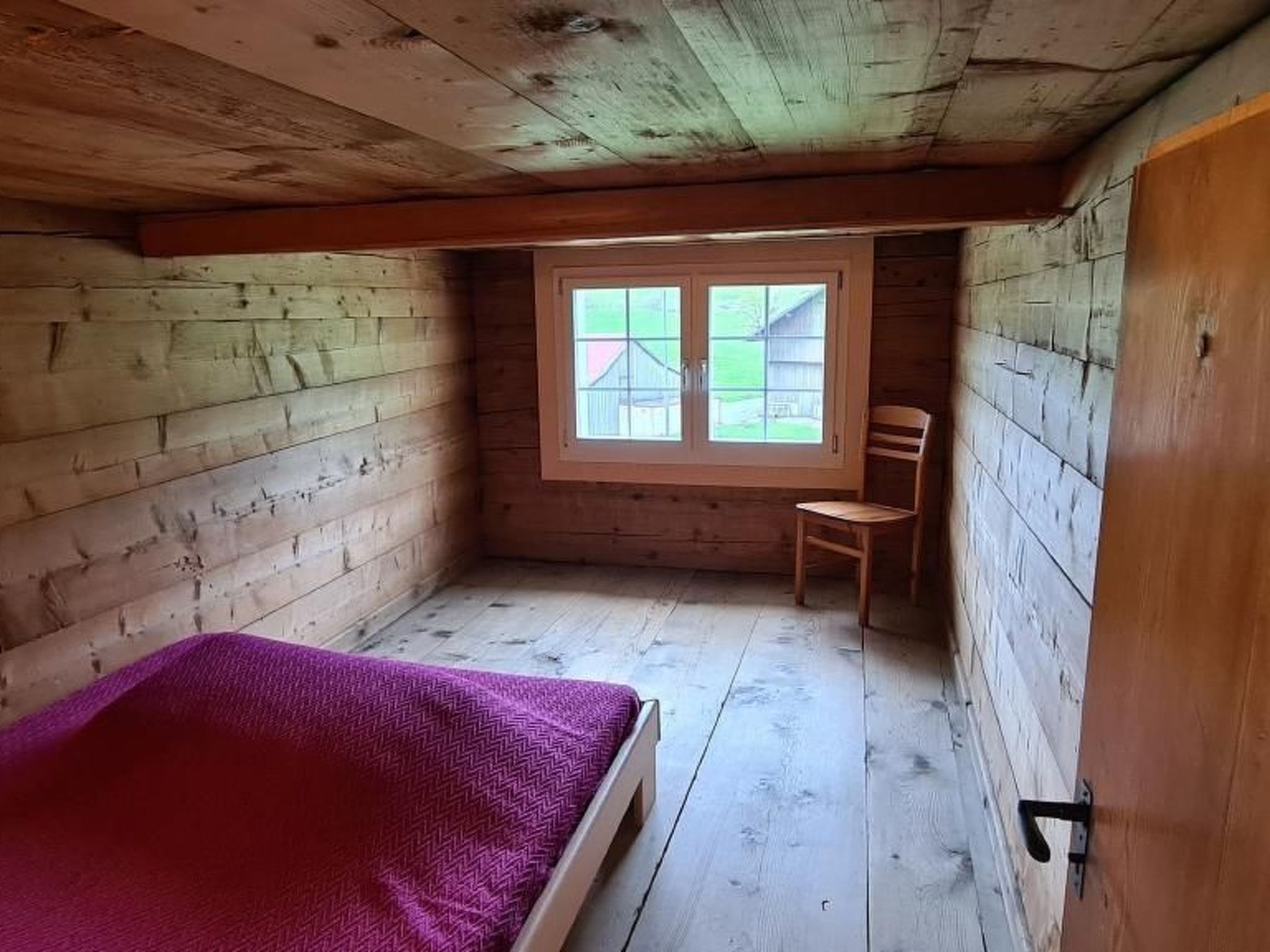 A small, wooden room with a single bed, a window, and a chair.