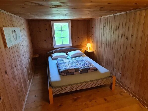 Una habitación pequeña y acogedora con paredes de madera y una cama individual. Hay una ventana que proporciona luz natural, y una mesita de noche con una lámpara junto a la cama.