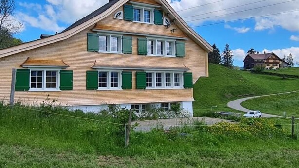 Ein zweistöckiges Haus mit grünen Fensterläden und einem geneigten Dach, umgeben von üppiger Vegetation und einer Serpentinenstraße.