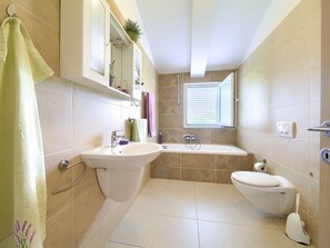 Ein Badezimmer mit Waschbecken, Toilette und Badewanne. Ein Fenster sorgt für natürliches Licht, und ein Handtuchhalter trägt ein aufgehängtes Handtuch.
