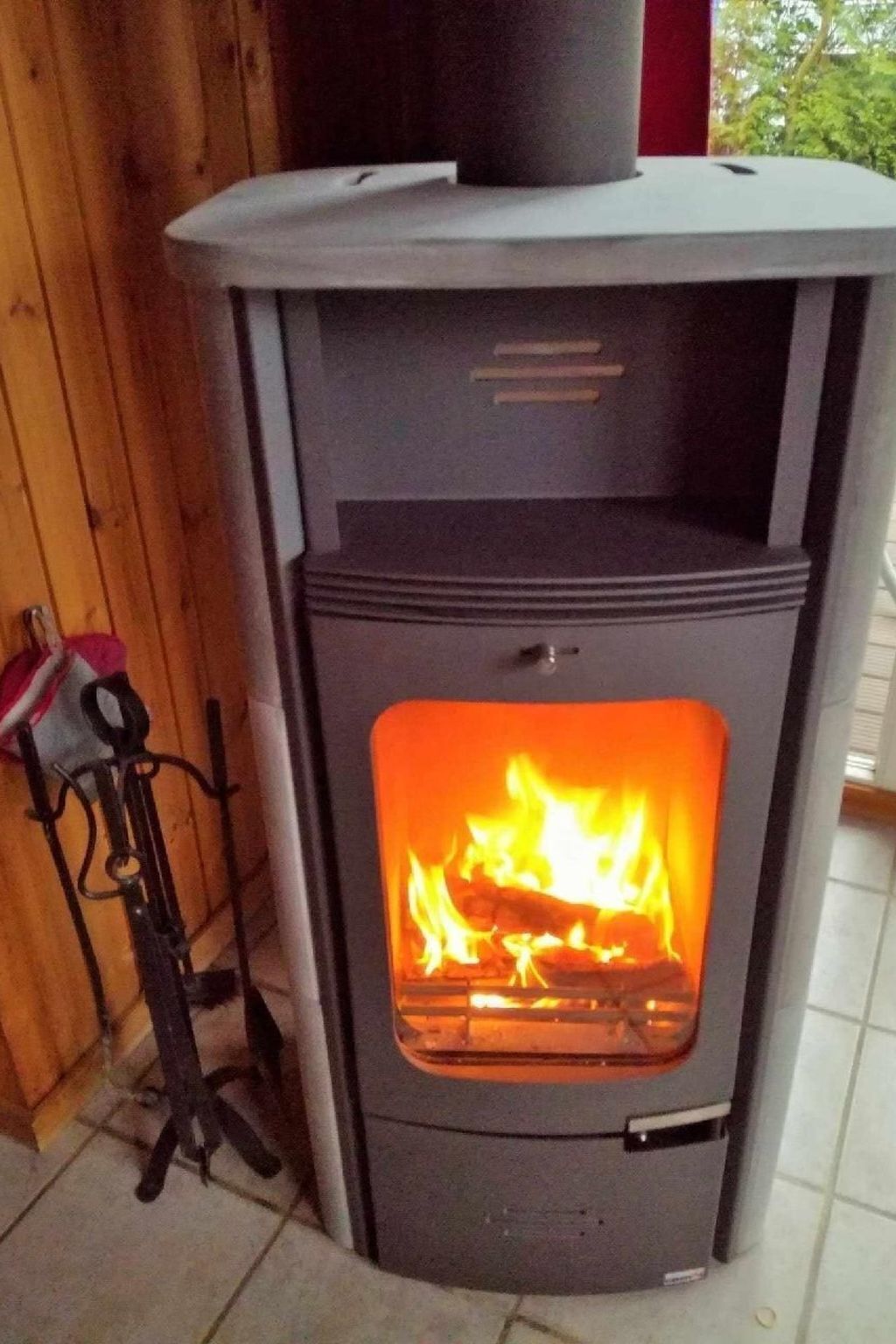 Un poêle à bois dont on peut voir les flammes à l’intérieur, un support en métal sur la gauche et un mur en bois en arrière-plan.