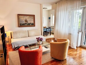 Ein Wohnzimmer mit einer Couch, einem roten Sessel, einem Couchtisch mit Blumenvase und einem Gemälde eines Schiffes an der Wand.