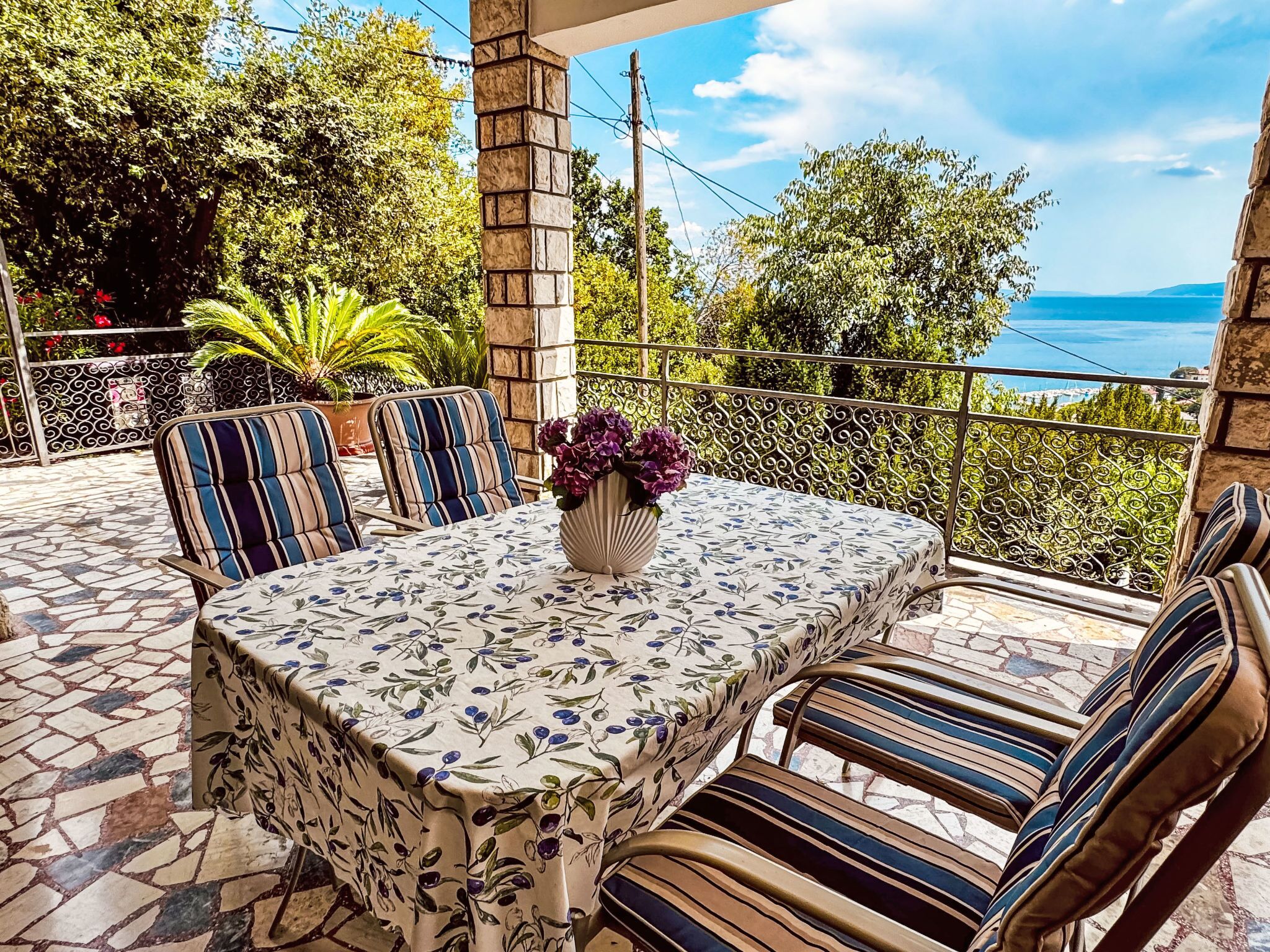 An outdoor patio with a table set for two, surrounded by striped chairs, a vase with flowers, and a view of the sea.