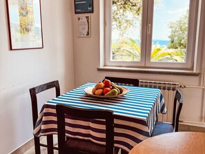 Ein Essbereich mit einem Tisch, der mit einer gestreiften Tischdecke bedeckt ist, Stühle, einer Obstplatte und einem Fenster mit Blick auf Grünflächen.
