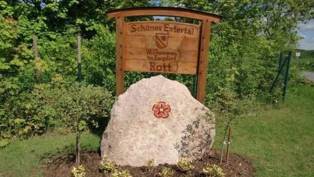 Ein hölzernes Schild mit Wappen und Schriftzug, umgeben von Grünpflanzen.