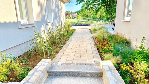 Ein gepflasterter Weg führt zu einem Gebäude mit Steinbord und einem Garten mit verschiedenen Pflanzen und Blumen.