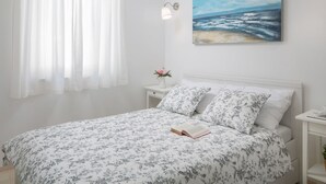 Ein Schlafzimmer mit einem Bett, einem Nachttisch, einer Wandleuchte, einem Strandbild und durchsichtigen Vorhängen.