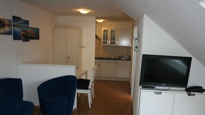 Ein Wohnzimmer mit Fernseher, zwei blauen Sesseln, einem kleinen Teppich und einer Küche im Hintergrund.
