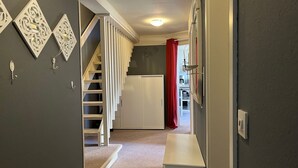 Un pasillo con piso alfombrado, una escalera con barandillas blancas y una cocina visible a través de una puerta abierta.