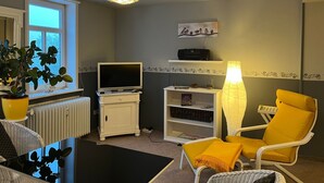Una sala de estar con televisión, estantería, sillón amarillo y una planta en maceta junto a la ventana.