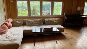 Ein gemütliches Zimmer mit Holzbalkendecke, einem großen Fenster mit Blick auf Grünflächen, einer Couch mit Kissen und einem kleinen Tisch mit Büchern.