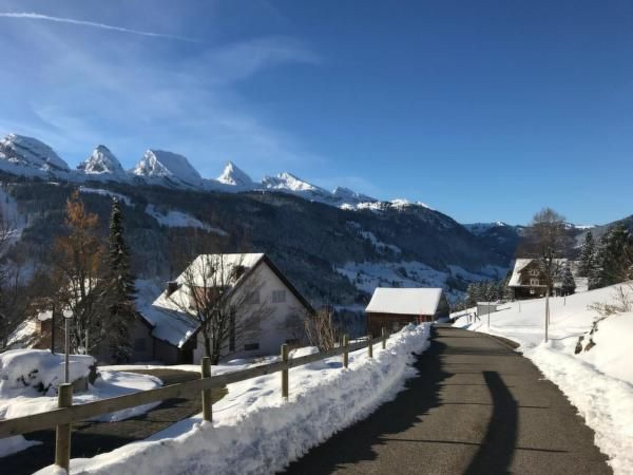 Carretera coberta de neu amb cases de fusta a banda i banda, que condueix cap a muntanyes nevades sota un cel serè.